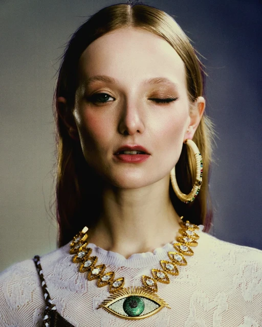 Close-up portrait of a woman with auburn hair wearing a white sweater, large hoop earrings, and a distinctive eye-shaped gold necklace.