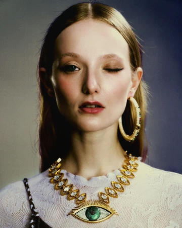 Close-up portrait of a woman with auburn hair wearing a white sweater, large hoop earrings, and a distinctive eye-shaped gold necklace.