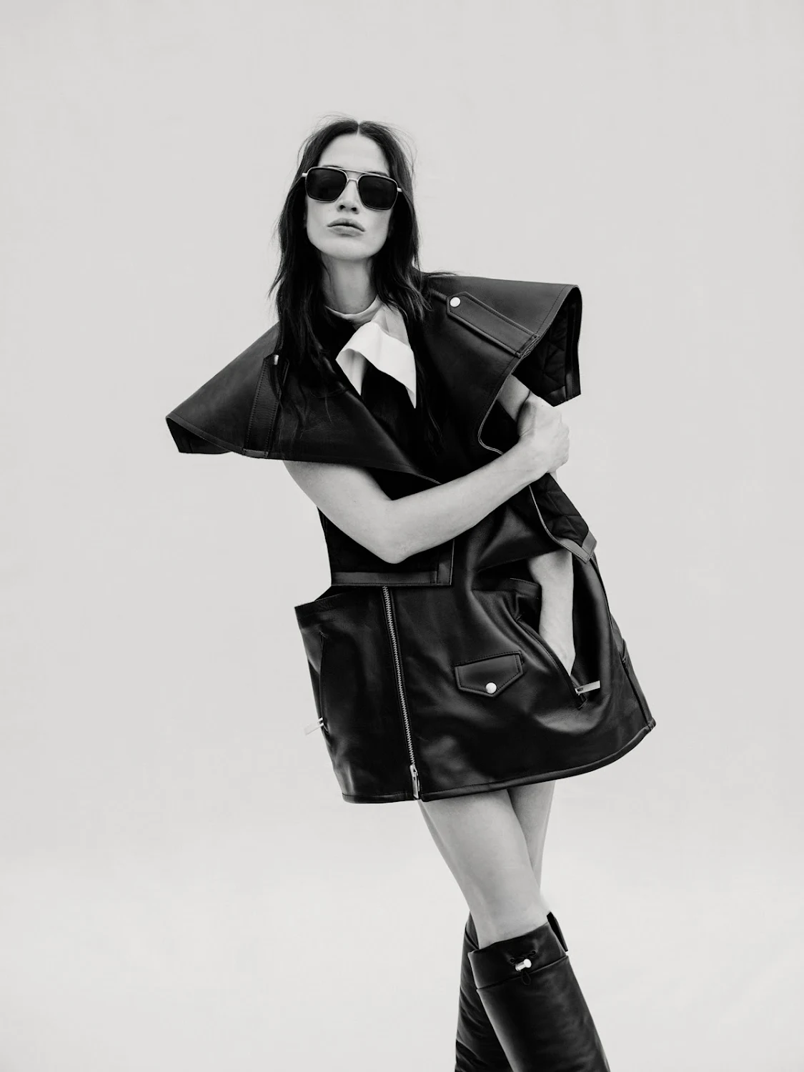 Monochrome fashion portrait of a woman in a black leather vest dress and knee-high boots, posing against a plain background.