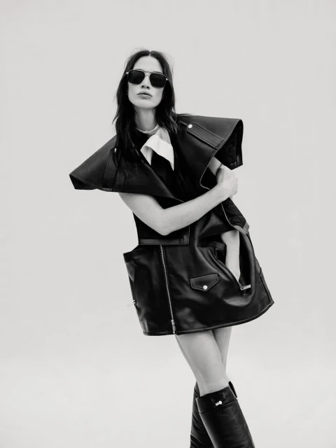 Monochrome fashion portrait of a woman in a black leather vest dress and knee-high boots, posing against a plain background.