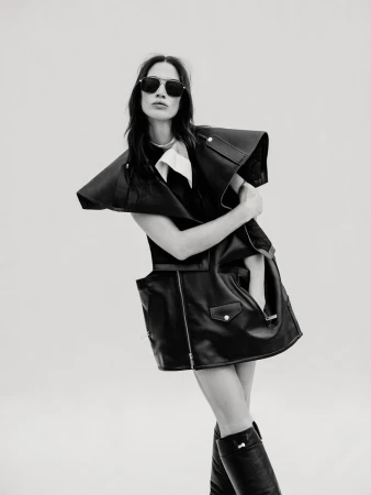 Monochrome fashion portrait of a woman in a black leather vest dress and knee-high boots, posing against a plain background.