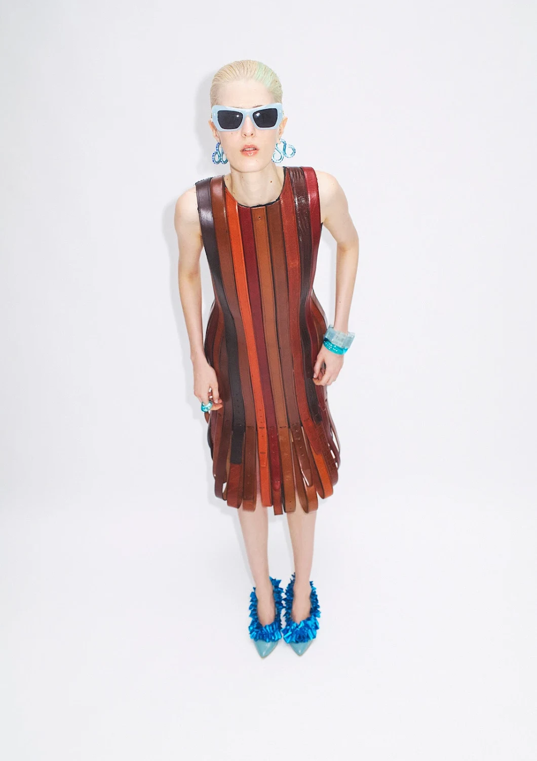Stylish young woman posing in a vertical-striped leather dress with bright-blue accessories and fuzzy heels against a white backdrop.