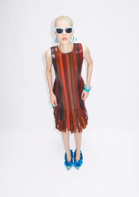 Stylish young woman posing in a vertical-striped leather dress with bright-blue accessories and fuzzy heels against a white backdrop.