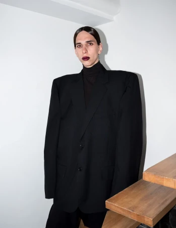 Model wearing an oversized black blazer and dark lipstick standing against a white wall near wooden stairs.