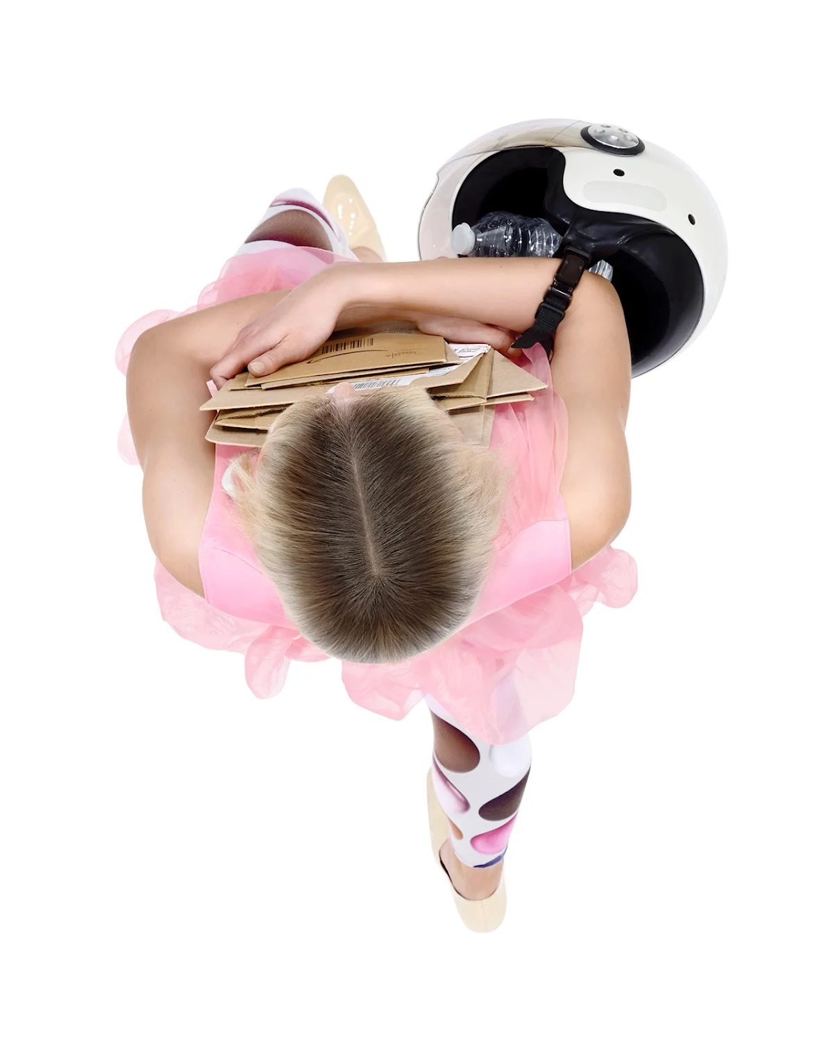 A young girl in a pink tutu, ballet shoes, and a motorcycle helmet embraces a stack of boxes, shot from an overhead perspective on white.