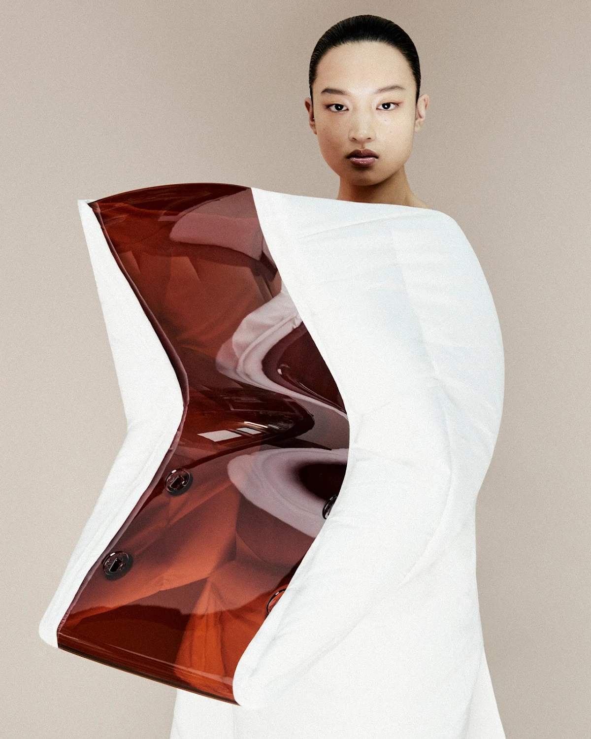A model in a structured white garment holding a large glossy curved brown object against a neutral background.
