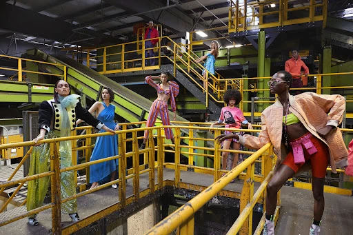 Multiple models in vibrant outfits pose amidst the industrial backdrop of a recycling plant, showcasing a fashion-forward editorial photoshoot.
