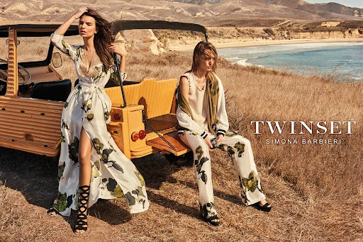 Two fashion models showcase TWINSET designs on a beach: one seated in cream pants, another in a floral dress next to a yellow vehicle.