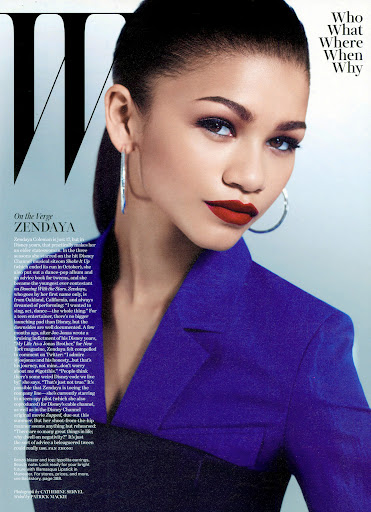Zendaya wearing a blue structured blazer and silver hoop earrings against a gradient background.