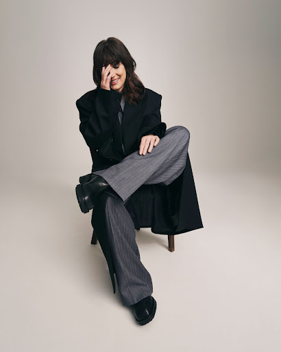 Portrait of a woman sitting on a stool, wearing a black coat and grey striped pants, covering her face with her hand indoors.