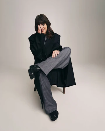 Portrait of a woman sitting on a stool, wearing a black coat and grey striped pants, covering her face with her hand indoors.