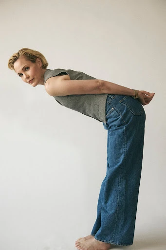 Adult female in a grey top and jeans bending backward, looking at the camera, and standing in front of a solid white background.