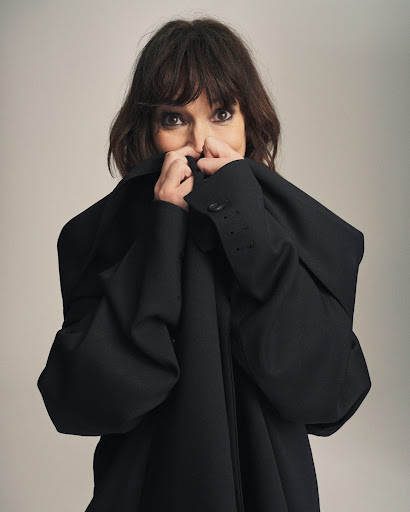 Portrait of an adult woman with brown hair and brown eyes, wearing a black jacket, holding the collar up to partially cover her face.