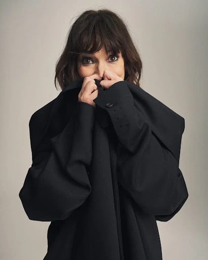 Portrait of an adult woman with brown hair and brown eyes, wearing a black jacket, holding the collar up to partially cover her face.