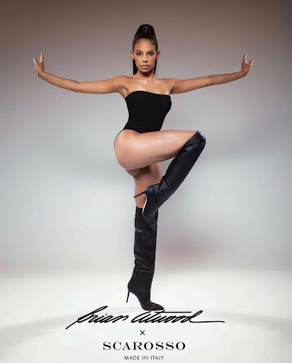 Woman in a black strapless bodysuit and knee-high leather boots posing against a neutral gradient backdrop