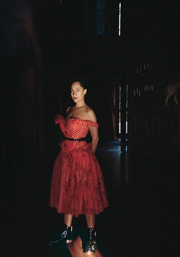 Woman in a red off-the-shoulder gown with ruffled details and black combat boots stands in a dimly lit, arched corridor.