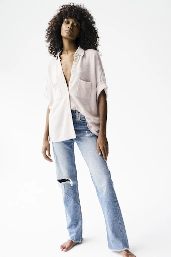 Full-length studio shot of a barefoot adult female model with curly hair wearing a light pink button-down shirt and light blue ripped jeans.