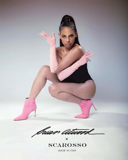 Fashion-forward shot of a woman in a black bodysuit, pink gloves, and pink heeled boots, posing in a studio against a grey background.