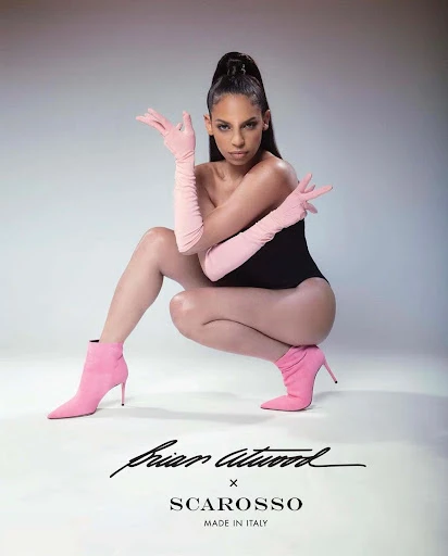 Fashion-forward shot of a woman in a black bodysuit, pink gloves, and pink heeled boots, posing in a studio against a grey background.