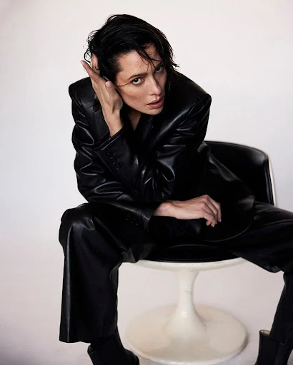 Adult female in a black leather suit poses while sitting in a white chair against a white backdrop in a studio setting.