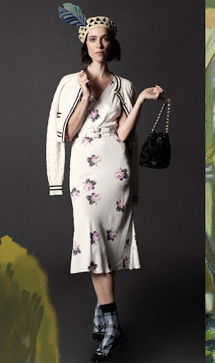 Elegant woman in floral dress, cardigan, and beret with feather, holding a Prada purse in a studio setting with painted backdrop.