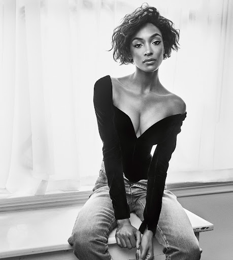 Black and white portrait of a young adult woman with short wavy hair, wearing a black top and jeans, seated by a window.