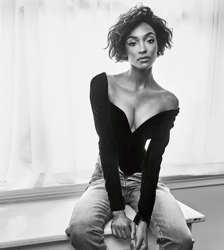 Black and white portrait of a young adult woman with short wavy hair, wearing a black top and jeans, seated by a window.