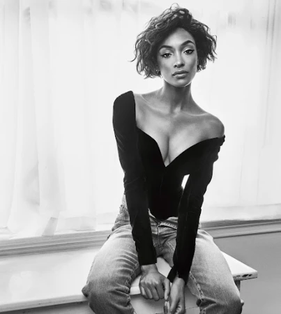 Black and white portrait of a young adult woman with short wavy hair, wearing a black top and jeans, seated by a window.