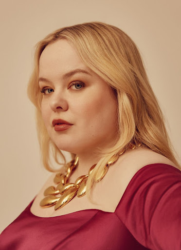 Portrait of a blonde woman in a burgundy dress and statement necklace looking at the camera in a studio setting.