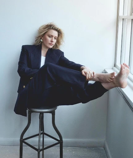 Portrait of a woman with blonde hair wearing a navy blue blazer and trousers, seated on a metal stool in front of a white wall.