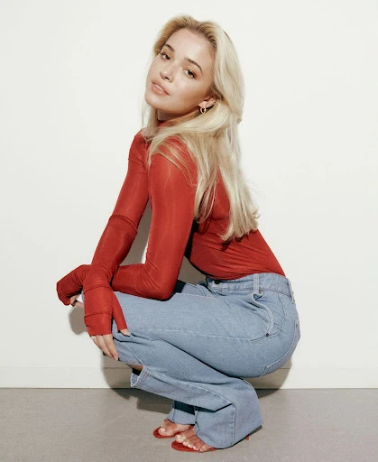 Blonde woman in a red sheer shirt and jeans squats in a photo studio, looking at the camera with a neutral expression.