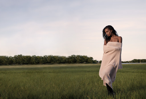 Woman wrapped in a beige blanket stands in a grassy field with a line of trees in the background under a light sky, outdoors.