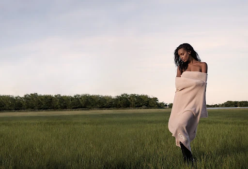 Woman wrapped in a beige knit blanket standing in a lush green meadow under an overcast sky.