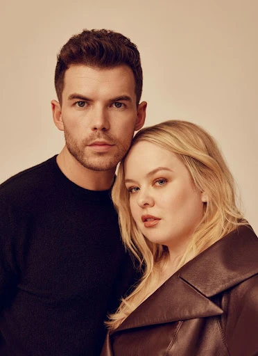 A portrait of a male and female couple, standing together, with the male wearing a black sweater and the female in a leather jacket.