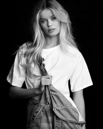 Monochrome portrait of a young woman with blonde hair, wearing a white t-shirt and holding denim overalls against a dark background.
