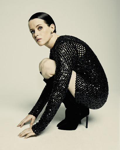 Elegant woman with short dark hair poses in a sequined mini dress and black boots against a neutral backdrop, looking at camera.