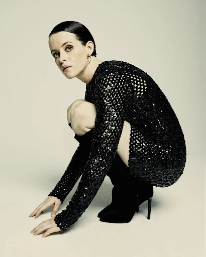 Elegant woman with short dark hair poses in a sequined mini dress and black boots against a neutral backdrop, looking at camera.