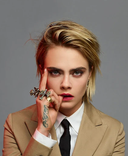 Close-up portrait of Cara Delevingne in a tan suit with finger to temple, showcasing blonde hair and a distinctive tattoo on her hand.