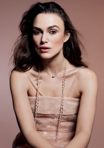 Portrait of Keira Knightley wearing a light pink dress with beaded necklaces, posing against a tan studio backdrop.