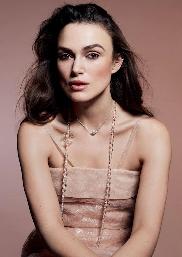 Portrait of Keira Knightley wearing a light pink dress with beaded necklaces, posing against a tan studio backdrop.