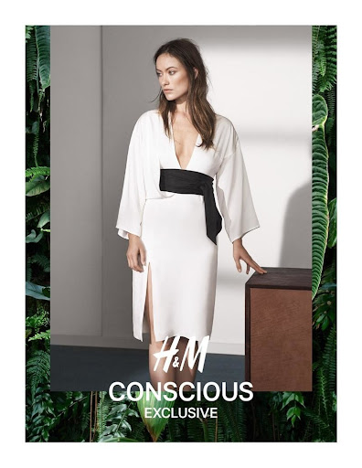 A fashionable woman in a white dress with a black belt poses in front of greenery and next to a brown prop. Fashion editorial.