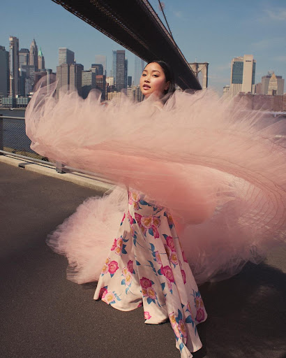 Woman poses in flowing pink tulle on the Brooklyn Bridge with the New York City skyline in the background on a sunny day.