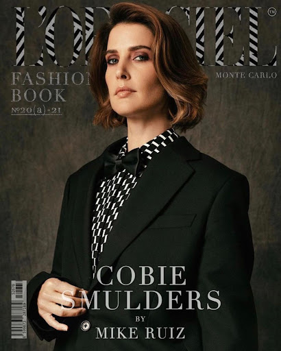 Cobie Smulders poses on a magazine cover wearing a black suit and black and white patterned shirt against a grey background.
