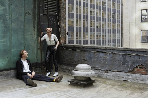 Two people pose on a rooftop with a cityscape in the background. A woman stands while a man sits on the roof of a building.