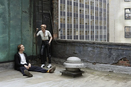 Two people pose on a rooftop with a cityscape in the background. A woman stands while a man sits on the roof of a building.