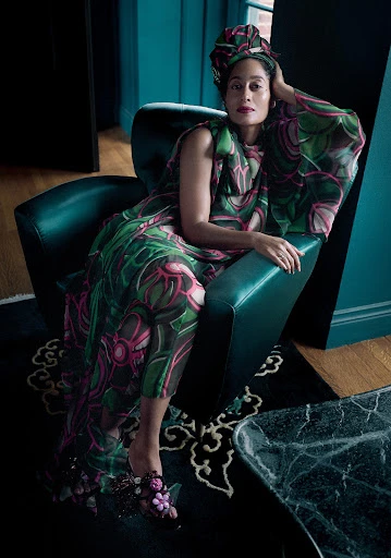 Woman wearing patterned dress and head wrap poses in a teal armchair on a black patterned rug indoors in a fashion editorial style.