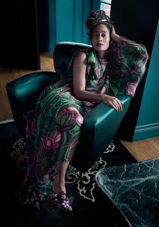 Woman wearing patterned dress and head wrap poses in a teal armchair on a black patterned rug indoors in a fashion editorial style.