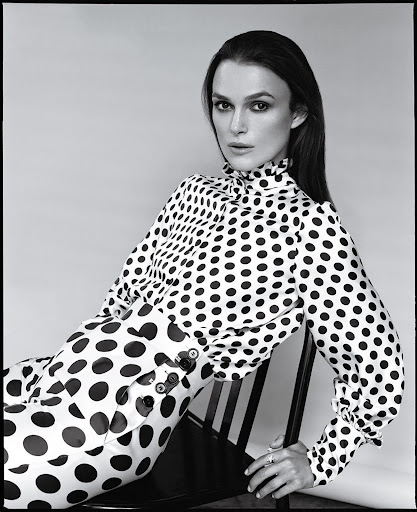 Black and white portrait of Keira Knightley wearing a polka dot top and pants while sitting on a black chair in a photo studio.