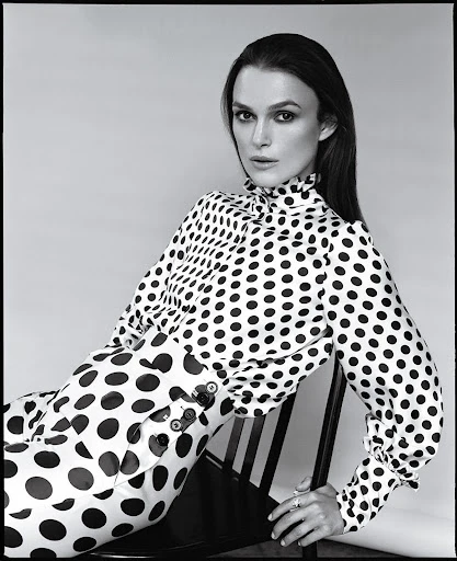 Black and white portrait of Keira Knightley wearing a polka dot top and pants while sitting on a black chair in a photo studio.