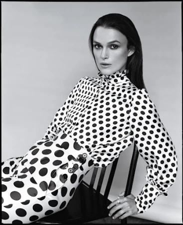 Black and white portrait of Keira Knightley wearing a polka dot top and pants while sitting on a black chair in a photo studio.